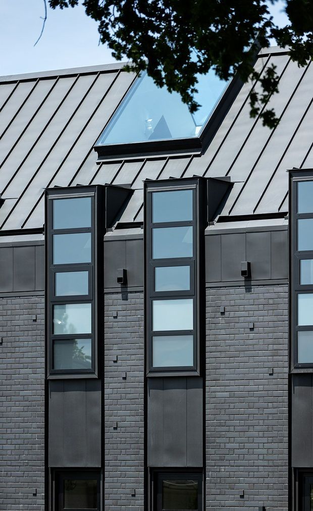 Close-up exterior view of a dark contemporary building facade with repeated narrow, vertically stacked windows and projecting dormer-like bays. The facade combines dark grey brickwork, dark metal cladding, black-framed glazing, and a steep standing-seam metal roof with a large angular skylight. Tree foliage partially frames the upper edge.