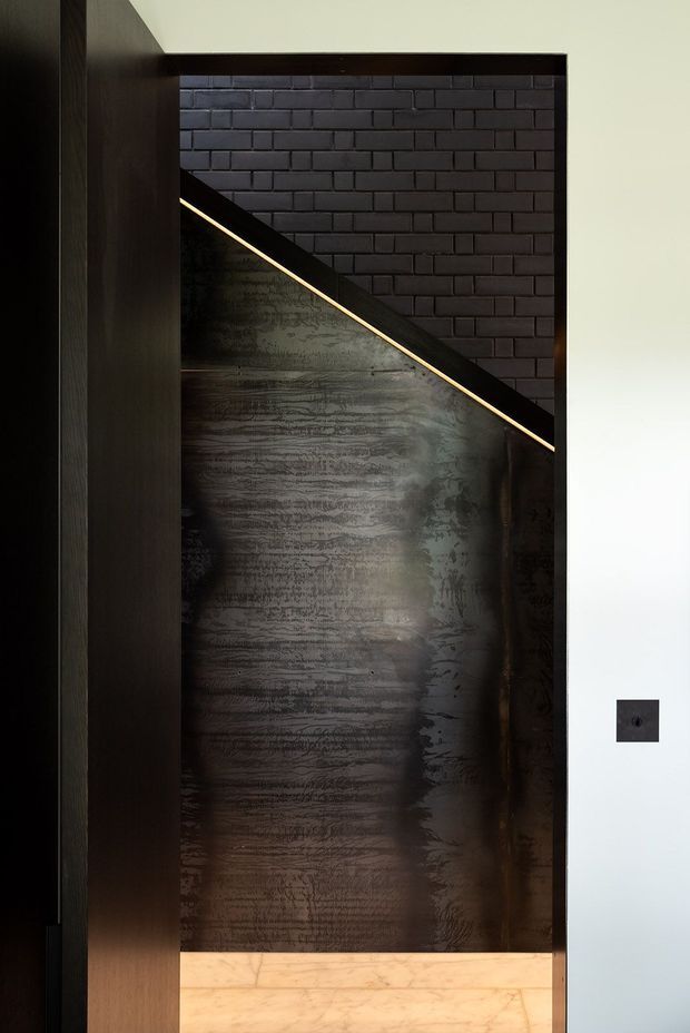 A close-up architectural interior detail showing a dark stair enclosure or feature wall. The composition combines black brickwork, a large dark textured metal panel, dark timber framing, and a slim linear LED light tracing the diagonal underside of the stair. A pale stone or marble floor and a white wall with a black switch provide contrast.