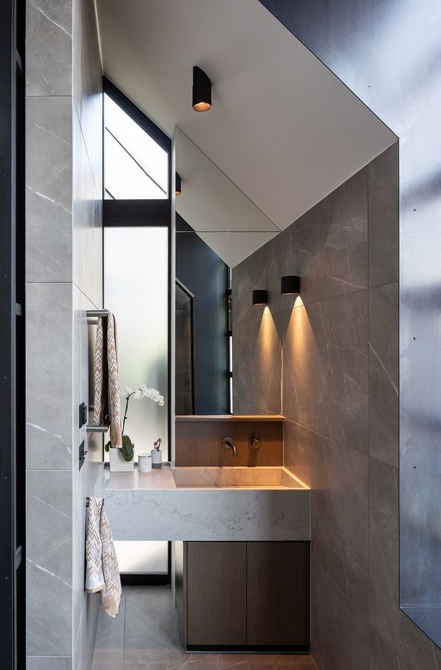 A narrow, contemporary bathroom with a floating dark timber vanity, pale stone benchtop and integrated basin, dark grey large-format tiled walls, and a full-height black-framed frosted window. A large mirror reflects the vanity area. Black wall sconces and a ceiling light provide warm accent lighting, while patterned towels and a small orchid add limited decoration.