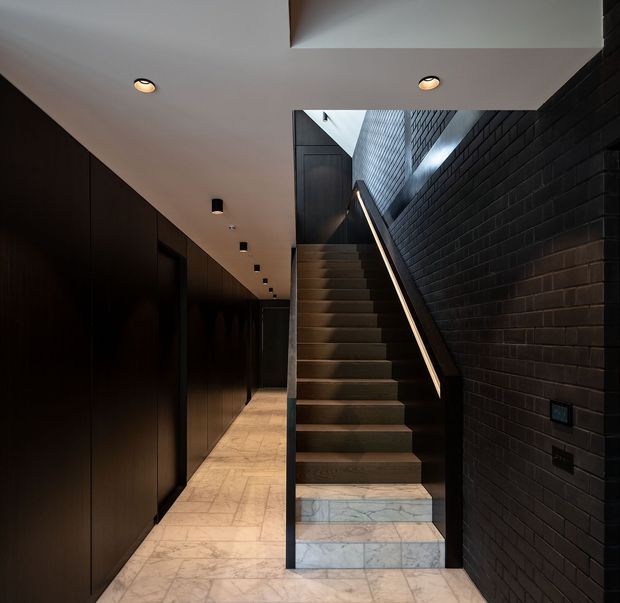 A dark, refined interior stairway and hallway. A straight timber staircase rises beside a black-painted brick feature wall, with a slim integrated handrail and a skylit upper landing. The adjacent corridor has full-height dark timber wall panelling, recessed ceiling lights, and pale veined stone floor tiles. The composition has strong linear geometry and a dramatic contrast between the dark walls and light flooring.