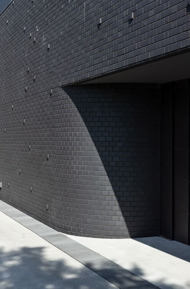 Close-up exterior architectural view of a dark charcoal brick facade with a recessed, shadowed entry or opening. The curved brick wall, crisp projecting canopy, dark doorway, and strong diagonal sunlight create a graphic, minimalist composition. A pale concrete and narrow grey paving strip run along the base.