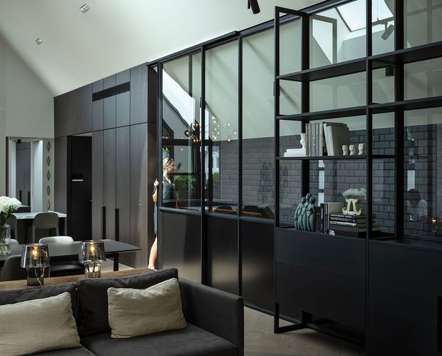 Open-plan contemporary interior with a dark, built-in kitchen and living area. A black-framed glass partition separates the spaces, while full-height dark cabinetry, dark grey brick-style wall tiles, black shelving, and charcoal upholstery create a moody, refined atmosphere. A dining table and chairs are visible to the left, with decorative objects and books arranged on the shelving.