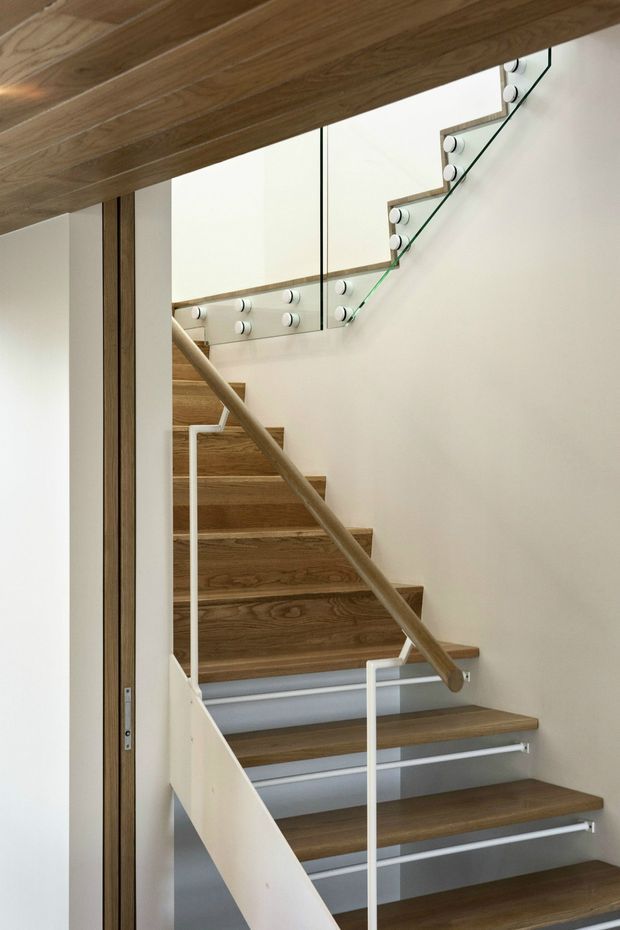 Interior view of a contemporary stairway with warm timber treads and soffit, white walls, slim white balustrades, and clear glass stair guards fixed with round metal fittings. A timber handrail runs diagonally through the stairwell, with a partial landing visible above.