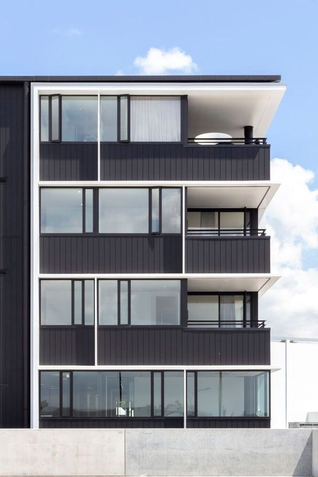 Exterior view of a contemporary multi-storey apartment building with a strong black-and-white facade composition. The building features repeated stacked balconies, dark vertical cladding, white projecting slab edges, extensive floor-to-ceiling glazing, dark metal window frames and balcony railings, and a flat roofline. A plain concrete boundary wall occupies the foreground under a bright blue sky.