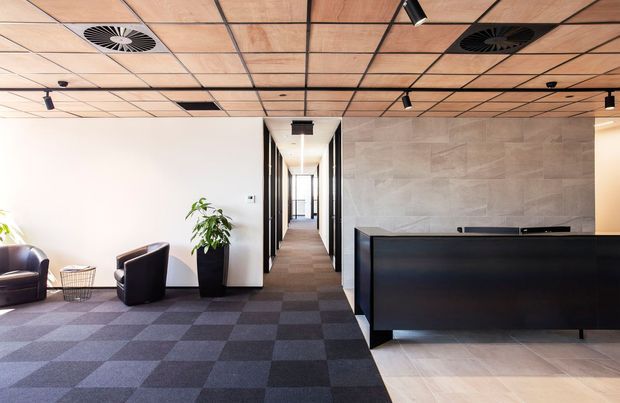 A contemporary commercial reception area with a large black reception desk, tiled feature wall, carpeted waiting zone, lounge chairs, and a corridor leading deeper into the office. The space has a suspended timber-toned acoustic ceiling grid with recessed services and track lighting, plus contrasting light timber-look flooring at the reception desk.