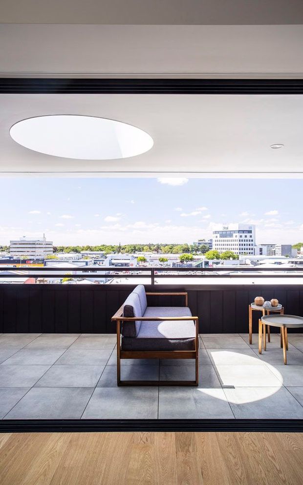 A bright contemporary balcony or terrace viewed from indoors through fully opened glazed doors. The terrace has large grey tiled flooring, a dark privacy balustrade, and mid-century-inspired outdoor lounge furniture including a timber-framed armchair and small side tables. A large circular roof opening or skylight above casts strong sunlight onto the terrace. Beyond the railing is a low-rise urban commercial landscape under a clear sky.