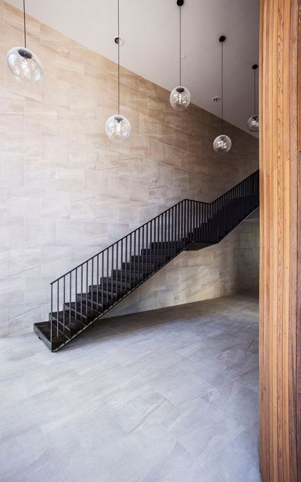 A tall, minimalist stair hall featuring a straight black metal staircase with slender vertical balusters, set against a full-height wall clad in large pale beige-grey stone or porcelain tiles. The floor continues in a light grey tiled finish. Several clear glass globe pendant lights hang at varied heights from a smooth light ceiling, while a vertical timber-clad wall or doorway frames the right edge. The space is restrained, contemporary, and softly lit.