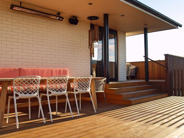A covered outdoor entertaining area with a timber deck, built-in red upholstered bench seating, a long dining table, and woven chairs. White brick walls, dark-framed glazing, timber steps, balustrades, and ceiling-mounted lighting define the space. The deck connects to an elevated entry or living area and appears designed for outdoor dining and relaxation.