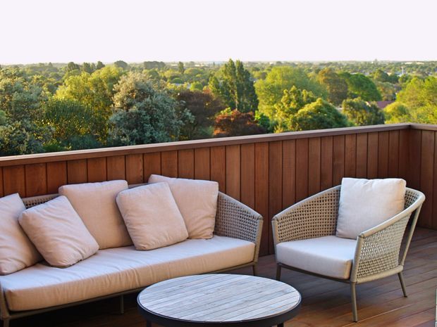 A furnished outdoor terrace or balcony with a cushioned woven sofa, matching armchair, and round slatted coffee table. A dark timber privacy balustrade frames expansive leafy suburban views in warm late-day light.