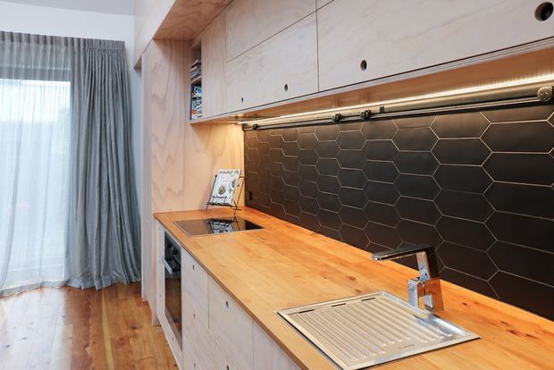 Compact contemporary kitchen with a long timber benchtop, integrated stainless-steel sink and drainer, pale timber plywood cabinetry, and a dark hexagonal tile splashback. Warm under-cabinet lighting, exposed timber flooring, and full-height grey curtains contribute to a practical, warm interior.