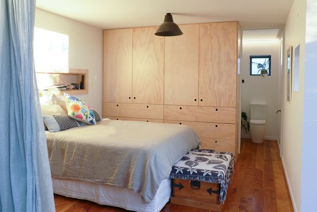 Bright, compact bedroom with a timber built-in wardrobe wall featuring tall panelled doors and multiple drawers. The room has warm timber flooring, a bed with light neutral bedding and colourful patterned cushions, a patterned upholstered bench at the foot of the bed, pale walls, a small wall niche, and a black pendant light. An open doorway leads directly to a small bathroom with a toilet visible.