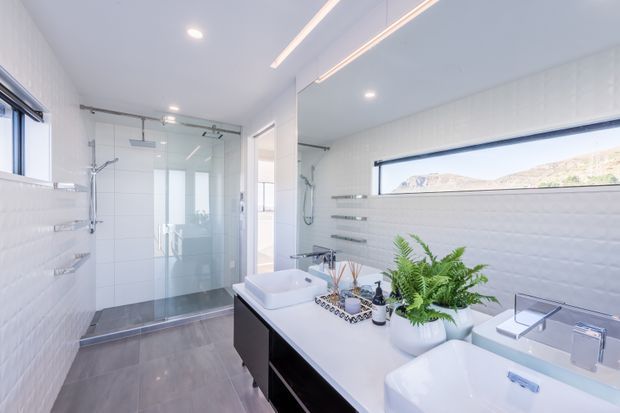 Bright, contemporary bathroom with a large double vanity, white vessel basins, dark cabinetry, white textured wall tiles, grey floor tiles, and a frameless glass walk-in shower. A long horizontal window provides natural light and views of the surrounding landscape.