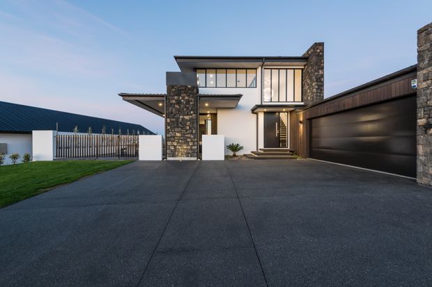 Contemporary two-storey residential facade photographed from the driveway at dusk. The home features a broad dark concrete driveway, a projecting flat-roof composition, extensive black-framed glazing, substantial stacked-stone wall elements, white rendered walls, and an attached dark double garage. A fenced lawn and neighbouring roofline are visible at the sides.