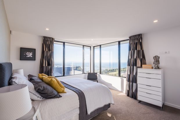 Bright, contemporary bedroom with a neatly made bed, upholstered bedside lamp, white drawer unit, patterned full-height curtains, and expansive corner glazing overlooking a coastal landscape. The room has neutral finishes, recessed ceiling lights, and soft grey and mustard bedding accents.