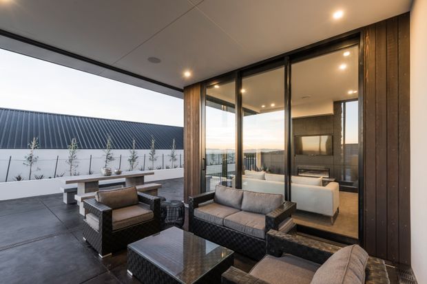 Contemporary outdoor lounge terrace adjoining a glazed living room, furnished with dark woven seating and a low reflective coffee table. The terrace has large-format dark paving, timber-clad structural columns, a raised planter with slender screening plants, and broad black-framed glazing opening to the interior. Inside, a warm neutral living area with a fireplace and built-in dark feature wall is visible through the glass.
