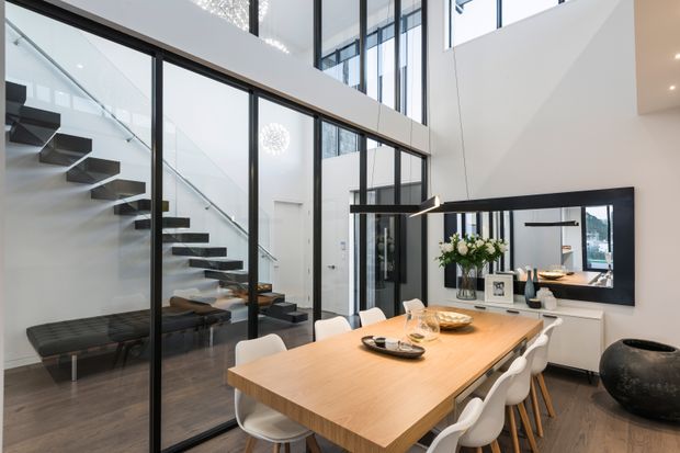 Bright contemporary dining area in a double-height interior, featuring a large timber dining table with white chairs, a black-framed glazed partition enclosing a floating staircase, dark timber flooring, and a large black-framed wall mirror. Floor-to-ceiling glazing and high-level windows create an airy, light-filled space.