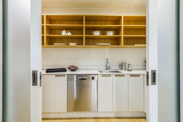 A compact, bright kitchen or scullery viewed through partially open sliding pocket doors. The space features pale built-in cabinetry, a stainless-steel dishwasher, white benchtops and splashback, open honey-toned timber shelving displaying crockery, and a sink beneath the shelving. Timber flooring and simple white architectural trim complete the clean, understated design.