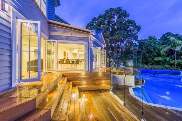 Twilight exterior view of a contemporary multi-level home opening onto a large timber deck with integrated steps and warm recessed lighting. Full-height glazed doors reveal a bright dining and kitchen area, while a glass balustrade borders a blue swimming pool surrounded by landscaped vegetation.