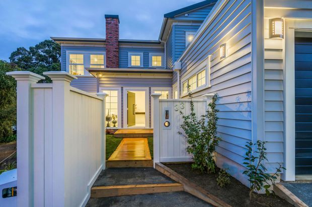 Twilight exterior view of a multi-level residential home approached via a narrow paved path and timber footbridge. The facade combines pale horizontal weatherboard cladding, white trim, a brick chimney, illuminated windows, a covered entry porch, and a white privacy fence and gate with planted borders.