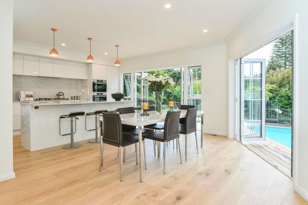 Bright open-plan kitchen and dining area with white built-in cabinetry, a central island with bar seating, pendant lights, pale timber flooring, and a dark dining table with leather chairs. Large glazed sliding and folding doors open directly to a pool area and landscaped garden, creating an airy indoor-outdoor connection.