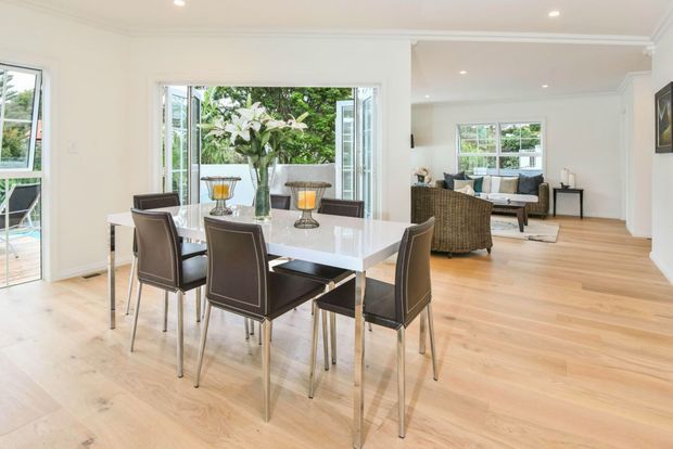 Bright open-plan dining and living area with a contemporary interior. A glossy white rectangular dining table with six dark upholstered chairs sits on light timber flooring, while the adjoining living room contains a sofa and woven armchairs. Large windows and glazed doors bring in abundant natural light and overlook greenery.