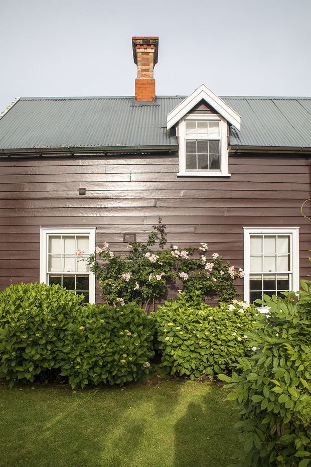 A front-facing exterior view of a compact traditional cottage with dark brown horizontal timber weatherboard cladding, a corrugated grey metal pitched roof, white-trimmed sash windows, a central brick chimney, and a small gabled dormer. Dense green hedges and flowering shrubs line the lawn in front.