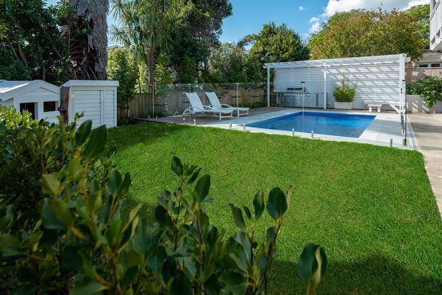 A landscaped backyard with a rectangular swimming pool, blue water, white pool coping, and a paved pool deck. White horizontal screening and a covered outdoor structure line the rear boundary, with two lounge chairs, potted plants, garden storage structures, mature trees, shrubs, and a broad manicured lawn creating a private residential outdoor setting.
