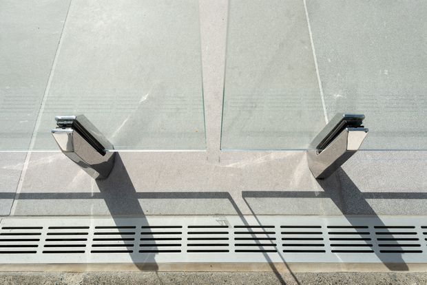 Close-up detail of a glass balustrade or barrier mounted on a light grey tiled or concrete surface. Two polished metal clamps or brackets secure the glass panels, with a long linear drainage grate running along the base. Strong sunlight creates sharp shadows.