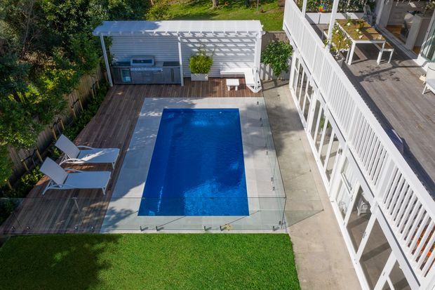Elevated view of a landscaped residential pool area with a rectangular blue swimming pool enclosed by clear glass fencing. A timber deck with two loungers runs alongside the pool, while a covered outdoor kitchen or entertaining area with a white pergola sits at the far end. The adjacent multi-level white-clad residence has broad timber decking and white balustrades, with lawn and mature greenery around the space.