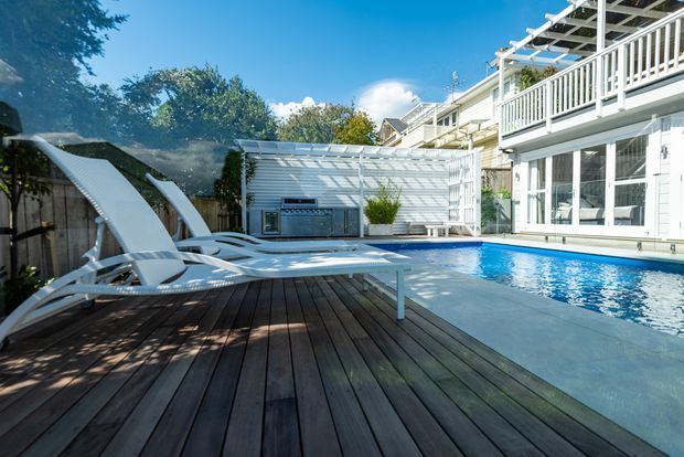 A bright residential pool area with a dark timber pool deck, white sun loungers, and a rectangular swimming pool beside a light-coloured multi-level house. White railings, glazed doors, pergola-like overhead framing, and a compact poolside outdoor entertaining structure are visible, surrounded by established greenery.