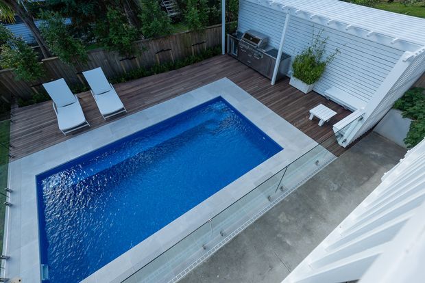 Elevated view of a compact rectangular swimming pool surrounded by pale paving and dark timber decking. The pool courtyard includes two lounge chairs, a built-in outdoor barbecue against a white horizontal-clad wall, potted greenery, and a glass safety balustrade.