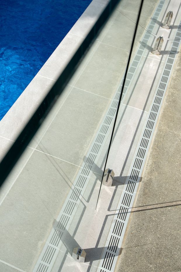 Close-up, top-down view of a swimming pool edge with pale tiled coping and pool surround. Long linear drainage grates run alongside the pool, with metal fixtures and strong geometric shadows visible on the light paving.