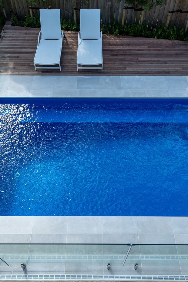 Top-down view of a vivid blue swimming pool bordered by pale stone or porcelain paving. A timber deck with two white reclining sun loungers sits along one edge, backed by a timber privacy fence and planting. Pool-edge drainage and a glass barrier are visible in the foreground.