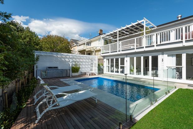 A bright, contemporary multi-level residential courtyard viewed from the garden, featuring a rectangular swimming pool enclosed by frameless glass safety fencing, a dark timber pool deck with loungers, a white rendered and weatherboard-style house, extensive white balcony railings, and a pergola over the upper terrace. A compact outdoor cooking area is set against the rear privacy screen.