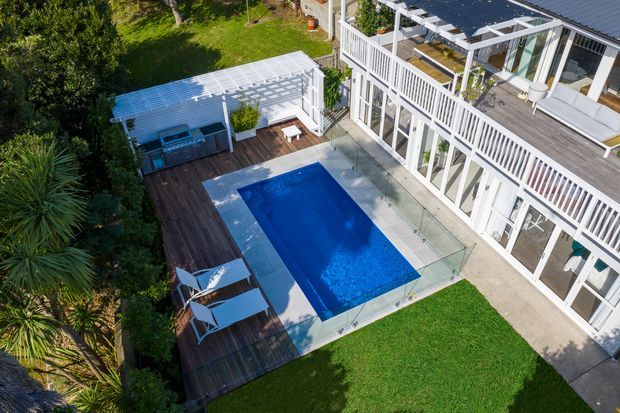 Aerial view of a landscaped residential outdoor area featuring a rectangular swimming pool, timber decking with sun loungers, a covered outdoor entertaining structure, and a white multi-level house with extensive glazed doors and balcony-style railings.