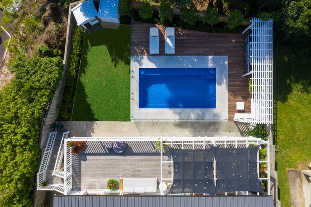 Aerial view of a landscaped residential outdoor area featuring a rectangular swimming pool, timber decking, concrete paving, a covered outdoor entertaining deck with shade sails, white pergola structures, lounge furniture, and surrounding lawn and planting.