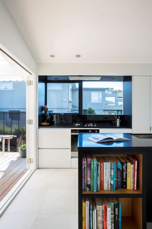 Bright contemporary kitchen viewed from an adjoining open doorway, featuring white flat-panel cabinetry, a glossy black splashback and benchtop, integrated appliances, and a dark island with open timber book shelving. Large windows provide natural light and overlook a small timber deck and neighbouring homes.