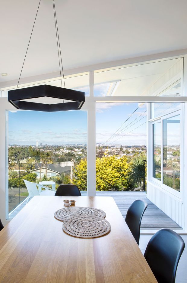 Bright dining area with a light timber dining table and black dining chairs, enclosed by large white-framed windows and an open glazed door. The room overlooks a leafy suburban landscape and distant city skyline. A geometric black pendant light hangs above the table, with a deck visible beyond the glazing.