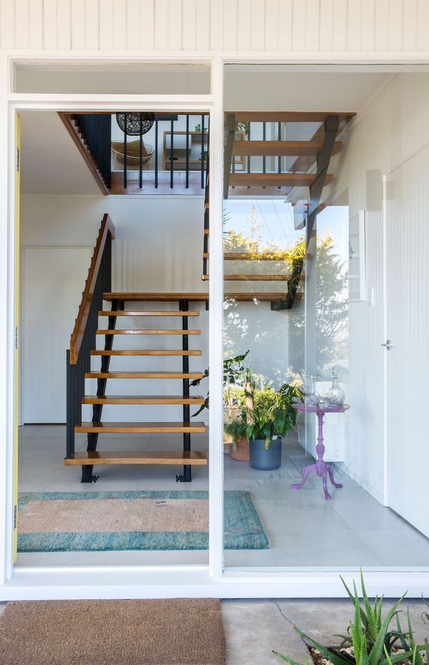 Bright architectural view into a contemporary entryway through large glazed doors. The space features an open-riser timber staircase with dark metal stringers and balustrade, white vertical wall panelling, pale tiled flooring, and a glazed upper landing. Potted plants, a small purple occasional table, and layered entry mats add colour and texture. The image also shows a sheltered exterior threshold and the home's facade glazing.