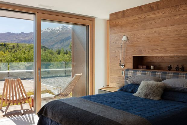 A warm modern bedroom with a large timber feature wall and built-in recessed bedside niche behind a neatly made bed. Floor-to-ceiling timber-framed glazing opens to a balcony or terrace with outdoor chairs, offering expansive views of green hills and snow-dusted mountains. Natural timber, dark blue bedding, and soft neutral textiles create a calm, comfortable retreat.