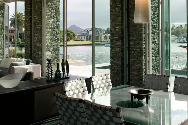 A refined waterfront dining area with a large glass dining table and woven armchairs, framed by floor-to-ceiling glazing and substantial stacked-stone columns. An adjacent living area is visible to the left, while a marina, landscaped waterfront homes, and boats appear outside. The interior has a strong connection to the water, with dark stone, glass, and neutral furnishings creating a contemporary, polished atmosphere.