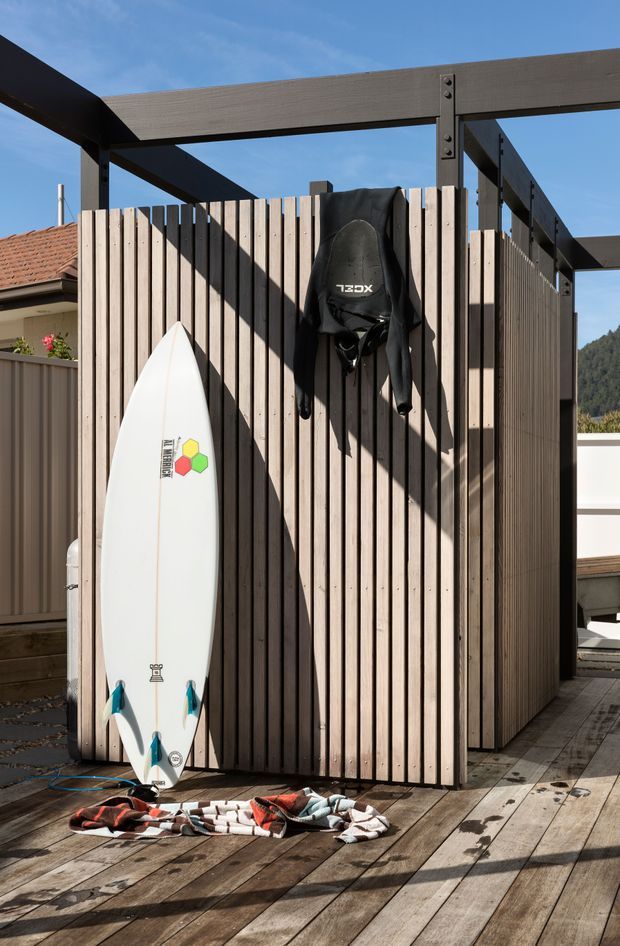 Outdoor timber-clad surf storage and changing enclosure on a raised timber deck, framed by dark exposed steel or timber beams. A surfboard and wetsuit hang against the vertical slatted wall, with strong sunlight and shadows creating a relaxed coastal outdoor setting.