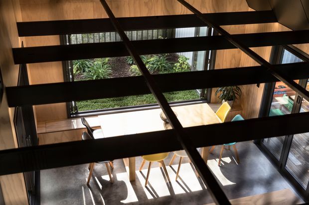 Elevated view into a contemporary dining area framed by exposed dark structural beams. The room features warm timber wall and ceiling surfaces, extensive windows overlooking greenery, a light dining table with colourful chairs, and a dark polished floor. Strong sunlight creates pronounced shadows across the space.