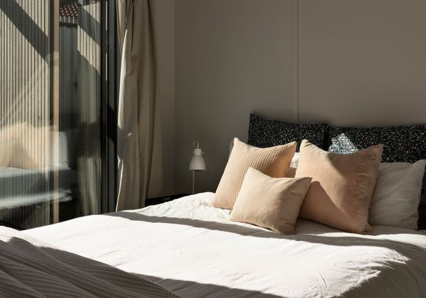 A sunlit contemporary bedroom with a neatly made bed, layered cream and beige pillows, dark patterned cushions, a neutral upholstered or plastered feature wall, and full-height glazed doors with sheer curtains. Strong diagonal sunlight creates a warm, calm atmosphere, with a small bedside lamp beside the bed.