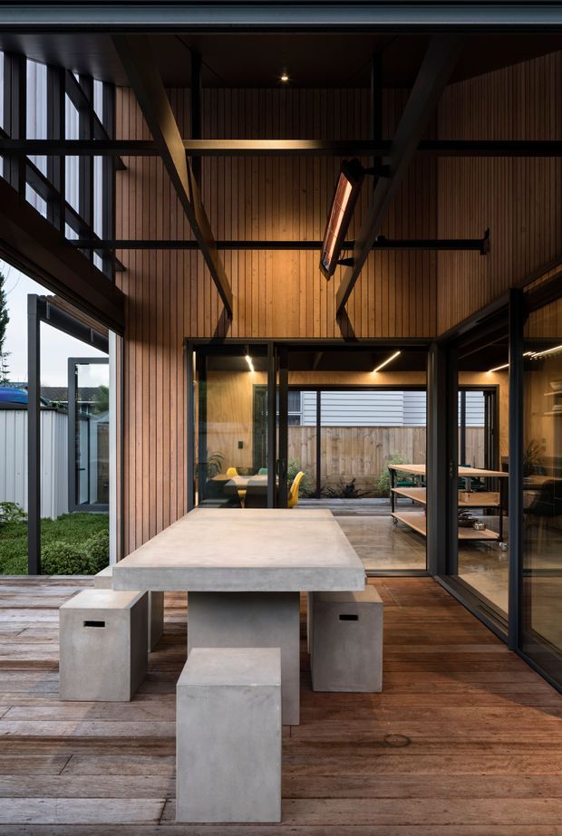 A covered outdoor dining terrace opening into a modern timber-clad pavilion. The space features a large concrete dining table with matching block stools, warm vertical timber wall lining, dark exposed steel framing, extensive black-framed glazing, timber decking, and built-in shelving. The open plan connects the deck to an adjacent interior lounge or work area and a planted courtyard.