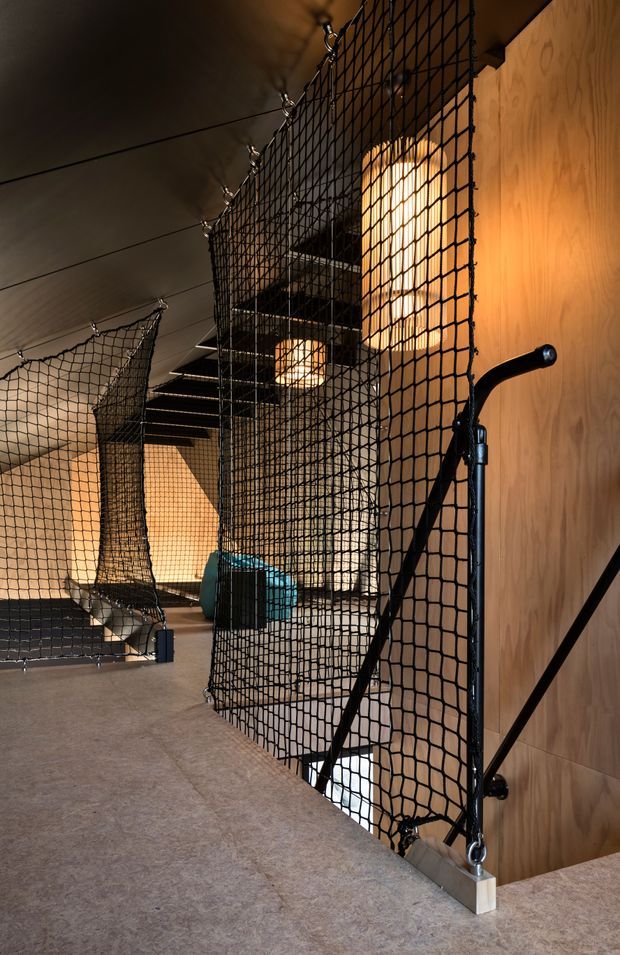 A dramatic interior circulation space with an industrial-style open staircase enclosed by black rope safety netting. Warm timber wall panelling, dark metal stair elements, exposed lighting, and a concrete-like floor create a utilitarian yet inviting multi-level environment.