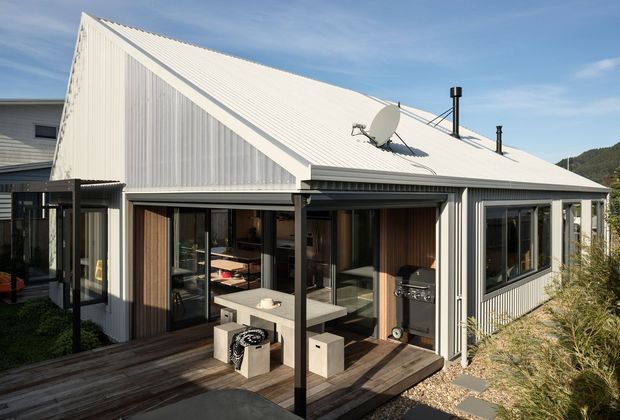 Exterior view of a contemporary single-storey home with a steep gabled corrugated metal roof and matching vertically ribbed metal cladding. Large glazed openings connect the interior to a timber deck and covered outdoor living area, with a compact outdoor grill and seating visible. The home sits on a sloping, vegetated site.