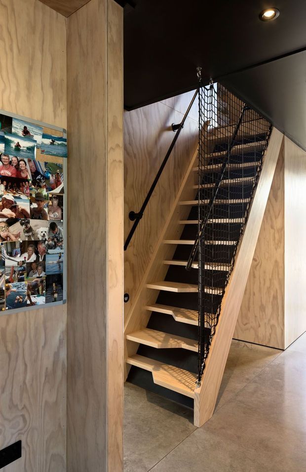 Interior view of a compact stairway with open timber treads, plywood-clad walls and a black metal mesh balustrade. The stair rises through a dark-painted ceiling, with black handrails and recessed lighting. A photo collage is mounted on the timber wall at left, and a concrete-look floor continues through the lower landing.