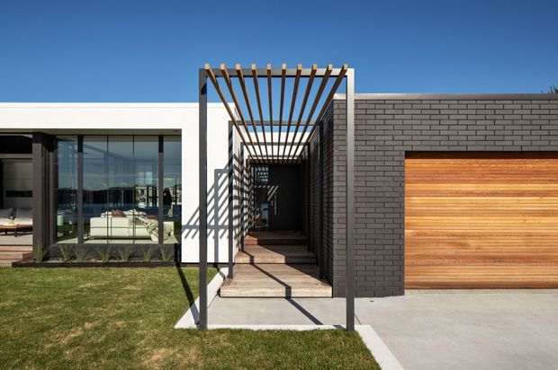 Contemporary single-storey house facade with a flat roof, dark grey brick garage wall, timber garage door, and expansive floor-to-ceiling glazing. A timber pergola creates a shaded covered entry walkway, with a neat lawn and concrete driveway in the foreground.