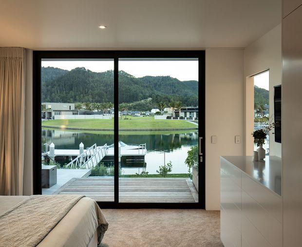 A refined bedroom with a large black-framed sliding glass door opening onto a timber deck and a tranquil waterfront marina. The room features soft neutral carpet, a partially visible upholstered bed, full-height curtains, recessed ceiling lighting, and sleek white built-in cabinetry. Forested hills and modern buildings are visible across the water.